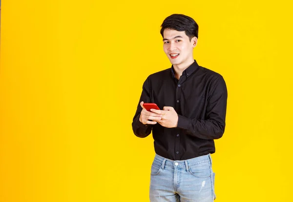 スタジオショットの千年のアジアの興奮男性ビジネスマンモデルでカジュアルな黒の長袖シャツとジーンズ立って手で赤いスマートフォンを保持黄色の背景に仮想ビデオゲームをプレイ — ストック写真