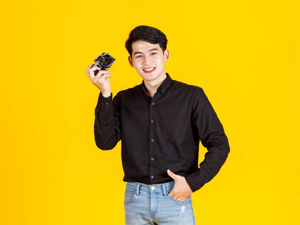 黄色の背景にデジタル一眼レフミラーレスカメラを目指して保持笑みを浮かべて立ってカジュアルな服装で千年のアジアの若いプロの男性ファッション写真家ジャーナリストのポートレートスタジオショット — ストック写真