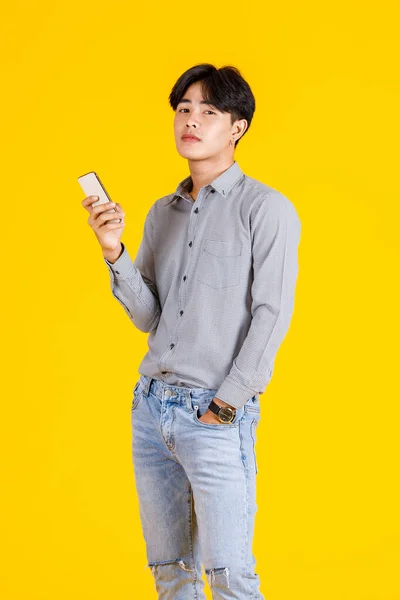 黄色の背景にスマートフォンを保持立ってスタイリッシュなファッショナブルなカジュアル衣装で千年のアジアの思慮深い疑わしい好奇心の男性ファッションモデルのスタジオショット — ストック写真