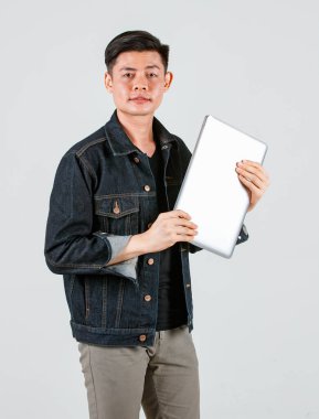 Asyalı yakışıklı erkek hippi modelin sıradan bir kot ceket giyip dizüstü bilgisayarı beyaz arka planda tutarken çekilmiş bir portre fotoğrafı.