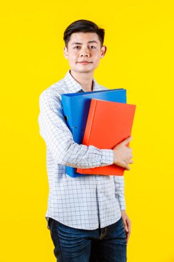 Asyalı genç iş adamının sıradan kıyafetleriyle çekilmiş bir portre resmi. Elinde evrak dosyası tutan kameraya bak. Sarı arka plandaki renkli dosyalar..