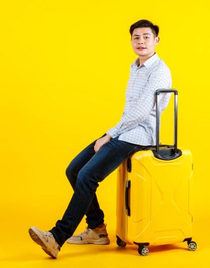 Milenyum Asyalı genç yakışıklı iş adamı modelinin portre fotoğrafı. Günlük kıyafet içinde. Büyük tramvay bagajında oturmuş gülümsüyor ve sarı arka planda iş gezisi için seyahat ediyor.