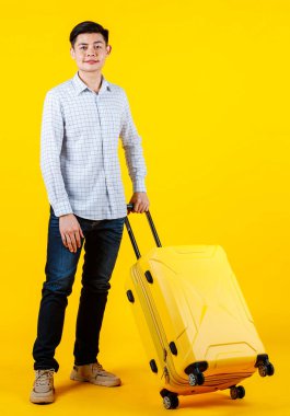 Asyalı genç ve yakışıklı bir iş adamının portre fotoğrafı. Günlük kıyafetlerle geziyor. Elinde büyük bir tramvay valizi, sarı arka planda iş seyahatine çıkarken gülümsüyor..