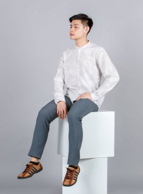 Portre stüdyosu, Asyalı mutlu, uzun kollu gömlekli, deri ayakkabılı, beyaz kare taburede oturan gri arka plandaki kameraya gülümseyen bir erkek model..