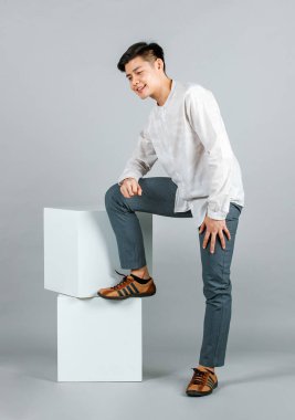 Portre stüdyosu, Asyalı mutlu, uzun kollu gömlekli, deri ayakkabılı, beyaz kare taburede oturan gri arka plandaki kameraya gülümseyen bir erkek model..