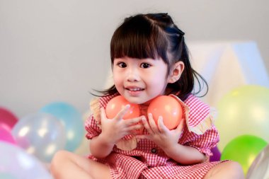 Küçük şirin anaokulu çocuğu, kırmızı uzun elbiseli, gülümseyen, gri arka planda renkli helyum hava balonlarıyla oynayan kız çocuğunun portresi..