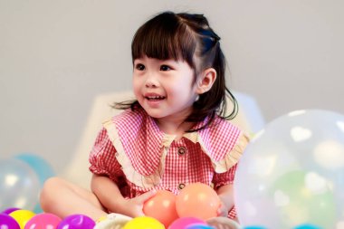 Küçük şirin anaokulu çocuğu, kırmızı uzun elbiseli, gülümseyen, gri arka planda renkli helyum hava balonlarıyla oynayan kız çocuğunun portresi..