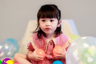 Küçük şirin anaokulu çocuğu, kırmızı uzun elbiseli, gülümseyen, gri arka planda renkli helyum hava balonlarıyla oynayan kız çocuğunun portresi..