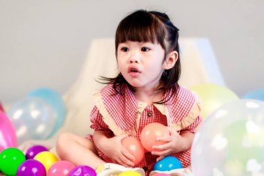 Küçük şirin anaokulu çocuğu, kırmızı uzun elbiseli, gülümseyen, gri arka planda renkli helyum hava balonlarıyla oynayan kız çocuğunun portresi..