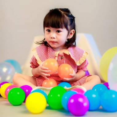 Küçük şirin anaokulu çocuğu, kırmızı uzun elbiseli, gülümseyen, gri arka planda renkli helyum hava balonlarıyla oynayan kız çocuğunun portresi..