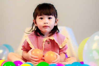 Küçük şirin anaokulu çocuğu, kırmızı uzun elbiseli, gülümseyen, gri arka planda renkli helyum hava balonlarıyla oynayan kız çocuğunun portresi..