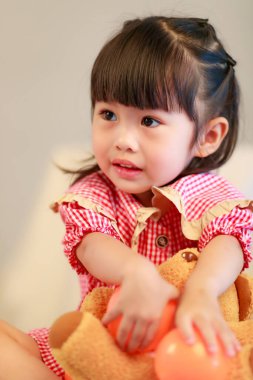 Küçük şirin anaokulu kızı kırmızı elbiseli, gülümseyen oyuncak ayıcığın gri arka planda renkli plastik toplarla oynarken çekilmiş bir portre..