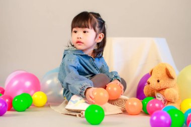 Küçük şirin anaokulu anaokulu kızı stüdyosu resmi. Günlük sokakta kot ceket giyer ve spor ayakkabı giyer. Elinde gri arka planda renkli plastik toplarla poz verir.