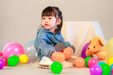 Küçük şirin anaokulu anaokulu kızı stüdyosu resmi. Günlük sokakta kot ceket giyer ve spor ayakkabı giyer. Elinde gri arka planda renkli plastik toplarla poz verir.