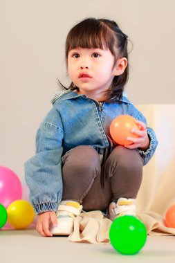 Küçük şirin anaokulu anaokulu kızı stüdyosu resmi. Günlük sokakta kot ceket giyer ve spor ayakkabı giyer. Elinde gri arka planda renkli plastik toplarla poz verir.