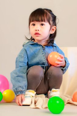 Küçük şirin anaokulu anaokulu kızı stüdyosu resmi. Günlük sokakta kot ceket giyer ve spor ayakkabı giyer. Elinde gri arka planda renkli plastik toplarla poz verir.