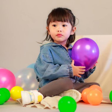 Gündelik sokakta oturan küçük tatlı anaokulu kızının atölyesi kot ceket giyer ve spor ayakkabı giyer. Yerde oturan gülümseyen oyuncak bebek gri arka planda renkli plastik toplarla oynar.