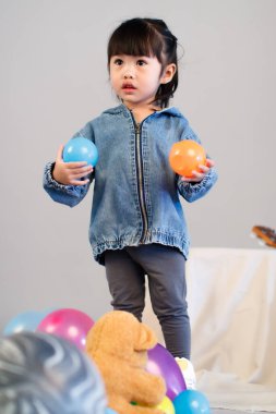 Stüdyoda, anaokulundaki küçük tatlı kız anaokulu çocuğunun tüm vücut fotoğrafları günlük sokakta kot ceket ve spor ayakkabıları giyer gri arka planda renkli plastik toplarla poz verir..