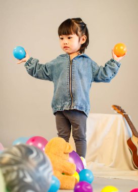 Stüdyoda, anaokulundaki küçük tatlı kız anaokulu çocuğunun tüm vücut fotoğrafları günlük sokakta kot ceket ve spor ayakkabıları giyer gri arka planda renkli plastik toplarla poz verir..
