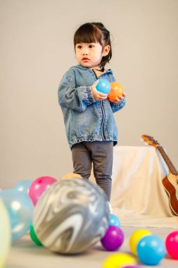 Stüdyoda, anaokulundaki küçük tatlı kız anaokulu çocuğunun tüm vücut fotoğrafları günlük sokakta kot ceket ve spor ayakkabıları giyer gri arka planda renkli plastik toplarla poz verir..