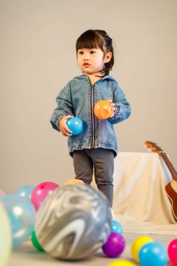 Stüdyoda, anaokulundaki küçük tatlı kız anaokulu çocuğunun tüm vücut fotoğrafları günlük sokakta kot ceket ve spor ayakkabıları giyer gri arka planda renkli plastik toplarla poz verir..