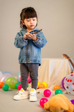 Küçük şirin, masum anaokulu anaokulu kızının resmi. Sıradan bir sokakta kot ceket giyiyor. Gri arka planda elinde akıllı telefonuyla kameraya bakıyor..