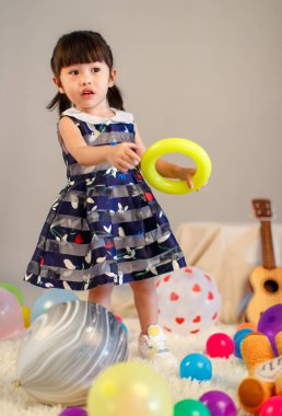 Küçük, masum anaokulu anaokulu kızı, mavi bir elbise giymiş, gri arka planda renkli balonlar tutan kameraya gülümseyen bir kızın portresi..