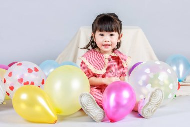Küçük şirin anaokulu çocuğu, kırmızı uzun elbiseli, gülümseyen, gri arka planda renkli helyum hava balonlarıyla oynayan kız çocuğunun portresi..