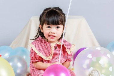 Küçük şirin anaokulu çocuğu, kırmızı uzun elbiseli, gülümseyen, gri arka planda renkli helyum hava balonlarıyla oynayan kız çocuğunun portresi..