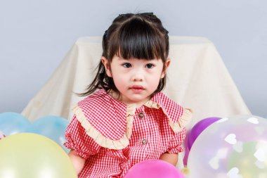 Küçük şirin anaokulu çocuğu, kırmızı uzun elbiseli, gülümseyen, gri arka planda renkli helyum hava balonlarıyla oynayan kız çocuğunun portresi..