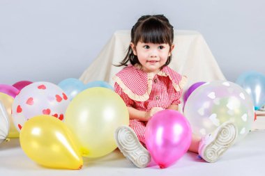 Küçük şirin anaokulu çocuğu, kırmızı uzun elbiseli, gülümseyen, gri arka planda renkli helyum hava balonlarıyla oynayan kız çocuğunun portresi..