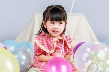 Küçük şirin anaokulu çocuğu, kırmızı uzun elbiseli, gülümseyen, gri arka planda renkli helyum hava balonlarıyla oynayan kız çocuğunun portresi..