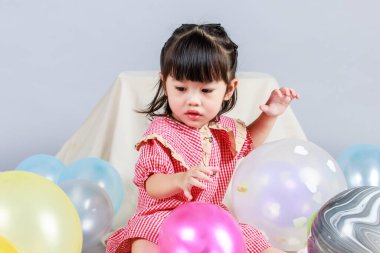 Küçük şirin anaokulu çocuğu, kırmızı uzun elbiseli, gülümseyen, gri arka planda renkli helyum hava balonlarıyla oynayan kız çocuğunun portresi..