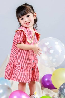 Küçük şirin anaokulu çocuğu, kırmızı uzun elbiseli, tek başına gülümseyen, gri arka planda renkli helyum hava partisi balonlarıyla oynayan kız çocuğunun portresi.