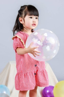 Küçük şirin anaokulu çocuğu, kırmızı uzun elbiseli, tek başına gülümseyen, gri arka planda renkli helyum hava partisi balonlarıyla oynayan kız çocuğunun portresi.