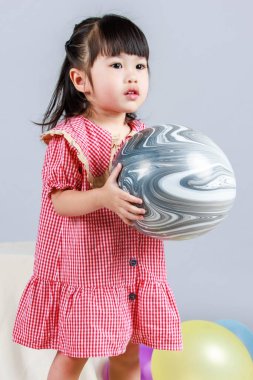 Küçük şirin anaokulu çocuğu, kırmızı uzun elbiseli, tek başına gülümseyen, gri arka planda renkli helyum hava partisi balonlarıyla oynayan kız çocuğunun portresi.