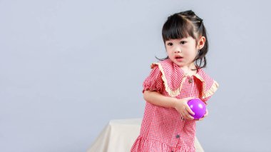 Küçük şirin anaokulu çocuğu, kırmızı uzun elbiseli, tek başına gülümseyen, gri arka planda renkli helyum hava partisi balonlarıyla oynayan kız çocuğunun portresi.