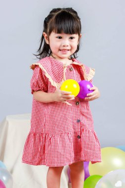 Küçük şirin anaokulu çocuğu, kırmızı uzun elbiseli, tek başına gülümseyen, gri arka planda renkli helyum hava partisi balonlarıyla oynayan kız çocuğunun portresi.