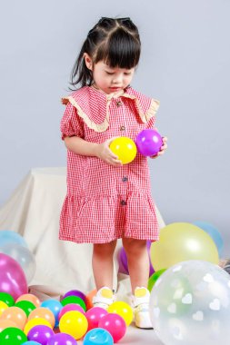 Küçük şirin anaokulu çocuğu, kırmızı uzun elbiseli, tek başına gülümseyen, gri arka planda renkli helyum hava partisi balonlarıyla oynayan kız çocuğunun portresi.