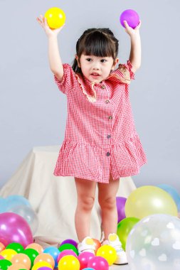 Küçük şirin anaokulu çocuğu, kırmızı uzun elbiseli, tek başına gülümseyen, gri arka planda renkli helyum hava partisi balonlarıyla oynayan kız çocuğunun portresi.