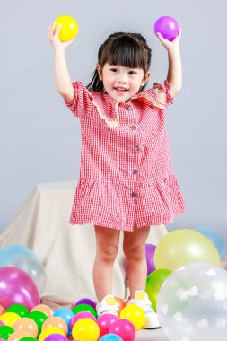 Küçük şirin anaokulu çocuğu, kırmızı uzun elbiseli, tek başına gülümseyen, gri arka planda renkli helyum hava partisi balonlarıyla oynayan kız çocuğunun portresi.