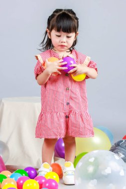 Küçük şirin anaokulu çocuğu, kırmızı uzun elbiseli, tek başına gülümseyen, gri arka planda renkli helyum hava partisi balonlarıyla oynayan kız çocuğunun portresi.