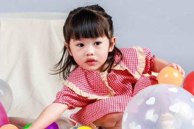 Küçük şirin anaokulu çocuğu, kırmızı uzun elbiseli, gülümseyen, gri arka planda renkli helyum hava balonlarıyla oynayan kız çocuğunun portresi..