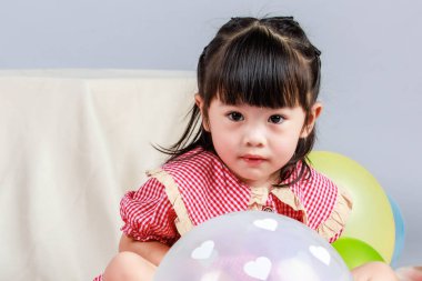 Küçük şirin anaokulu çocuğu, kırmızı uzun elbiseli, gülümseyen, gri arka planda renkli helyum hava balonlarıyla oynayan kız çocuğunun portresi..
