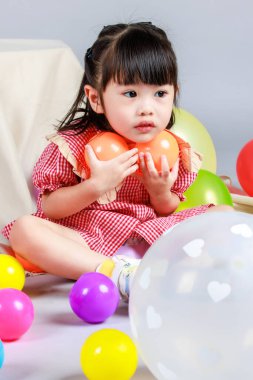 Küçük şirin anaokulu çocuğu, kırmızı uzun elbiseli, gülümseyen, gri arka planda renkli helyum hava balonlarıyla oynayan kız çocuğunun portresi..