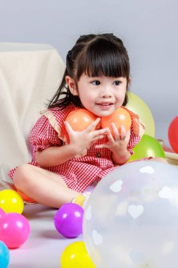 Küçük şirin anaokulu çocuğu, kırmızı uzun elbiseli, gülümseyen, gri arka planda renkli helyum hava balonlarıyla oynayan kız çocuğunun portresi..
