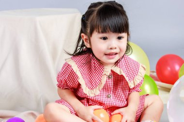 Küçük şirin anaokulu çocuğu, kırmızı uzun elbiseli, gülümseyen, gri arka planda renkli helyum hava balonlarıyla oynayan kız çocuğunun portresi..