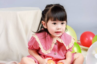 Küçük şirin anaokulu çocuğu, kırmızı uzun elbiseli, gülümseyen, gri arka planda renkli helyum hava balonlarıyla oynayan kız çocuğunun portresi..