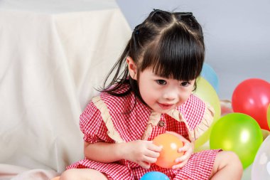 Küçük şirin anaokulu çocuğu, kırmızı uzun elbiseli, gülümseyen, gri arka planda renkli helyum hava balonlarıyla oynayan kız çocuğunun portresi..