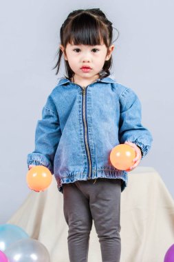 Stüdyoda, anaokulundaki küçük tatlı kız anaokulu çocuğunun tüm vücut fotoğrafları günlük sokakta kot ceket ve spor ayakkabıları giyer gri arka planda renkli plastik toplarla poz verir..